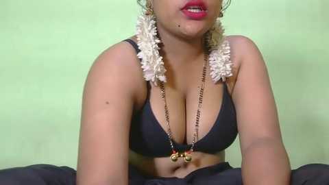 Video of a light-skinned woman with medium-sized breasts, wearing a black bra with gold chains, and white flower garland, against a pale green background.