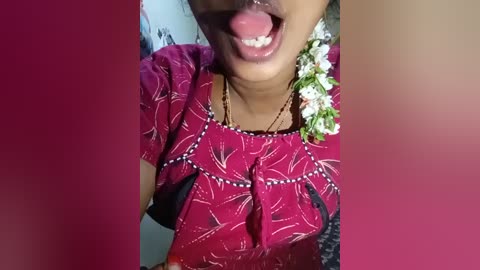 Video of a woman with dark skin, wearing a maroon blouse with white leaf patterns, and a white flower in her hair. She is smiling with her tongue out. Background is blurred.