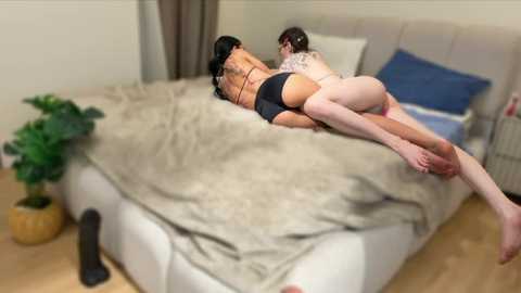 A video captures two women, one with tattoos, lying closely on a beige bed with a cream blanket, in a cozy, dimly lit room.