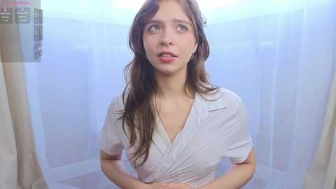 Video of a young woman with long brown hair, wearing a wet, white, V-neck shirt, standing in a dimly lit, tiled shower stall with translucent walls, looking surprised and slightly distressed.