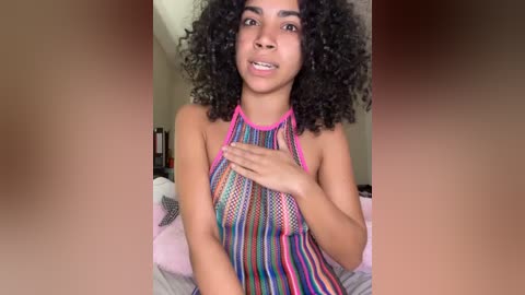 Video of a young Black woman with curly hair, wearing a colorful striped halter top, sitting on a bed with a pink blanket, smiling, and covering her chest.