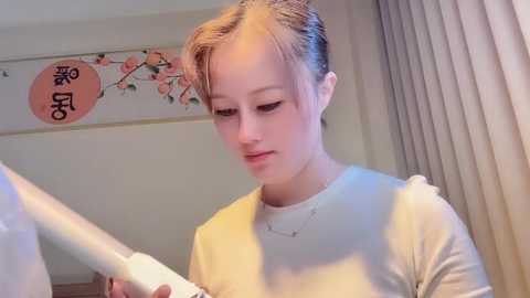 A video of a young, fair-skinned woman with blonde hair in a ponytail, wearing a white shirt, examining a white object in her hands. Background includes a beige curtain and Japanese decorative banner with cherry blossoms.