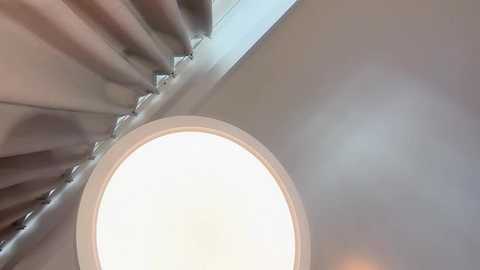 Video of a modern room with a cylindrical ceiling light fixture emitting soft white light. The ceiling is beige, and there are vertical, metallic blinds partially drawn to the right. The image is taken from a low angle, focusing on the light and blinds.