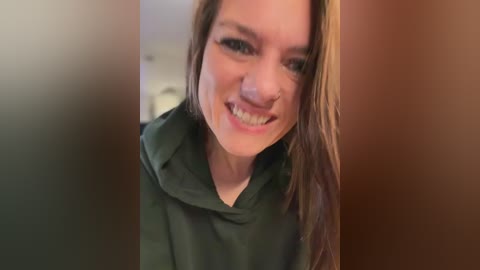A video of a smiling, fair-skinned woman with straight, shoulder-length brown hair, wearing a dark green hoodie, standing in a blurred, warm-toned indoor hallway.