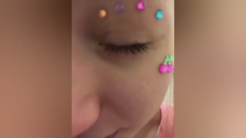 Video of a young woman with closed eyes, adorned with colorful rhinestone eye decorations in shades of blue, green, pink, and yellow.