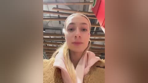 Media: A video of a young blonde woman with fair skin, wearing a pink bathrobe, taken in a cluttered room with wooden beams and a red garment hanging in the background.