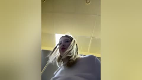 Video of a woman with long blonde hair, wearing a white sweater, captured from a low angle inside a room with beige walls and a fluorescent ceiling light.