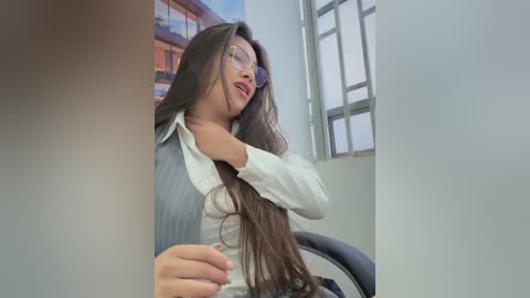 Video of a young woman with long dark hair, wearing glasses, a white blouse, and a gray vest, sitting in an office with a window and a colorful mural background.