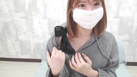 Video of an East Asian woman with short brown hair and a white mask, wearing a gray hoodie, adjusting a black shoulder strap, sitting on a light blue chair against a white, patterned wall.