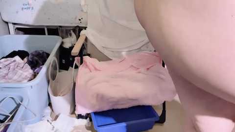 A video of a nude person, possibly a woman, wearing pink lingerie, standing in a cluttered bathroom with a white sink, blue basket, and scattered clothes.