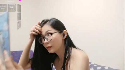 A video of an East Asian woman with long black hair and glasses, wearing a black top, adjusting her hair in a bathroom with a white wall and blue shower curtain.