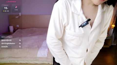 A video shows a woman in a white blouse and black skirt, leaning over a bed with a white duvet in a dimly lit bedroom.