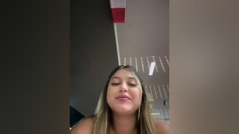 A video of a young woman with light skin, long blonde hair, and closed eyes, possibly in a bathroom, with a tiled wall and red accents visible in the background.