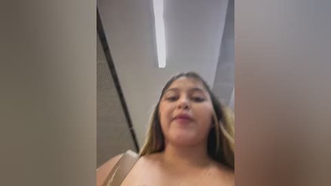 Video of a young woman with light brown skin and long, straight, blonde hair, standing in a dimly lit, modern bathroom with white walls and a ceiling light. She is topless, with visible breasts.