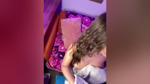 Video of a young woman with long, wavy brown hair, topless, sitting on a bed with a colorful patterned blanket, a cushion, and a wooden frame in the background.