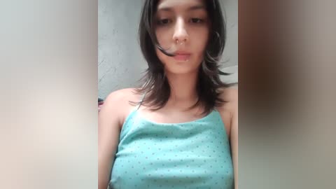 Video of a young woman with medium skin tone, shoulder-length dark hair, wearing a light blue spaghetti-strap tank top. She has a slim build, and the background is a plain, light-colored wall.