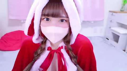 Video of a young woman with light skin, wearing a red and white Santa costume with a white face mask, sitting on a white floor, in a room with pink curtains and white furniture.