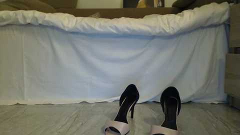 Video of a person lying on a beige carpeted floor, wearing black high heels with white soles, with white bedding draped over their legs, suggesting a moment of rest.