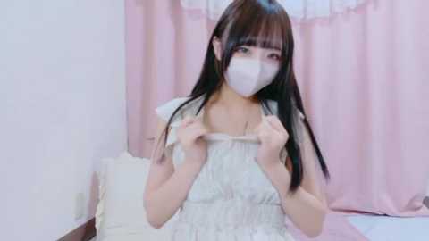 Media: A video of an East Asian woman with long black hair, wearing a white surgical mask and a sleeveless, white, ruffled dress. She stands in a room with pastel pink curtains and white bedding, holding her dress straps.
