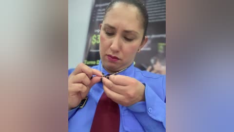 Video of a woman with light skin and brown hair tied back, wearing a blue button-up shirt and maroon top, adjusting a necklace in a classroom setting with a blackboard and blurred figures in the background.