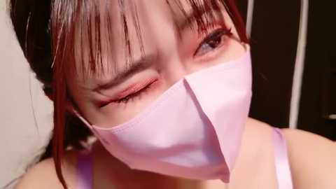 Close-up video of an Asian woman with straight, shoulder-length dark hair, wearing a light pink surgical mask, winking, and showcasing a subtle, delicate makeup look. Background is blurred, with a hint of a dark wall.