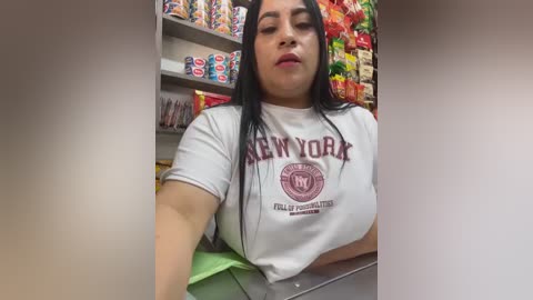 Video of a Latina woman with long black hair, wearing a white New York Knicks t-shirt, working at a candy store with colorful, assorted candy displays in the background.