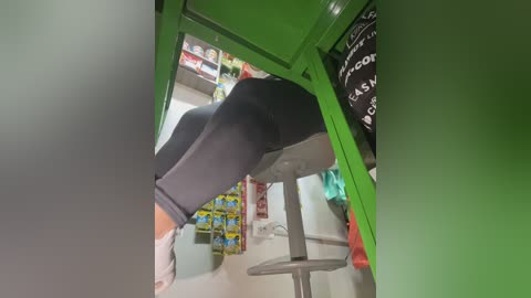 A video capturing a person wearing black leggings and a light-colored shirt, leaning over a green shelf, with colorful snacks and a black bag visible in the background.