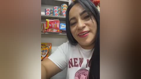 Video of a smiling Latina woman with long black hair, wearing a white New York Yankees t-shirt, standing in a well-lit convenience store. Shelves behind her display colorful snack packages.