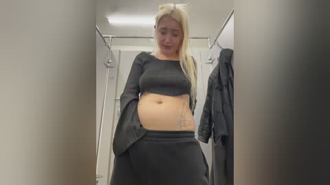 Video of a pregnant, blonde woman with fair skin in a changing room. She wears a black crop top and matching pants, revealing a tattoo on her side.
