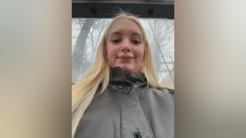 Video of a smiling, fair-skinned blonde woman with long hair, wearing a gray jacket, standing in front of a window with a blurred, wintry outdoor scene visible behind her.