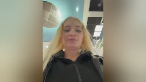 Video of a blonde Caucasian woman with long hair, wearing a black hoodie, taken from a low angle, showing her from the chest up, in a modern, brightly lit room with blue walls and circular light fixtures.