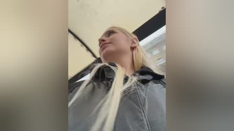 Video of a blonde woman with fair skin, wearing a black jacket and a white scarf, looking upward, with a blurred background of an indoor setting.