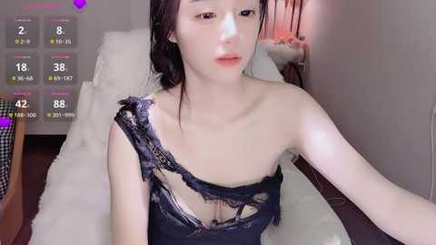 Video of a slender Asian woman with fair skin and black hair, wearing a torn black lace top, lying on a white bed in a dimly lit room.