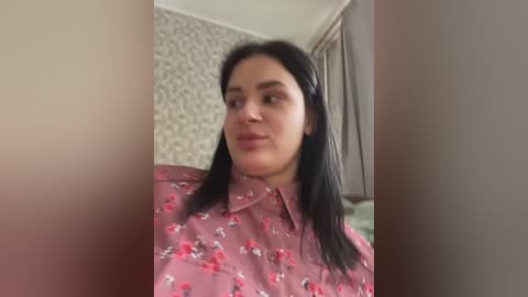 Video of a middle-aged woman with straight black hair, wearing a pink floral blouse, standing in a room with patterned wallpaper and a wooden door.