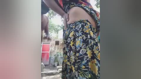A video shows a woman in a floral-patterned skirt, lifting her top to reveal her midriff, in a garden setting with a red door and green potted plants in the background.