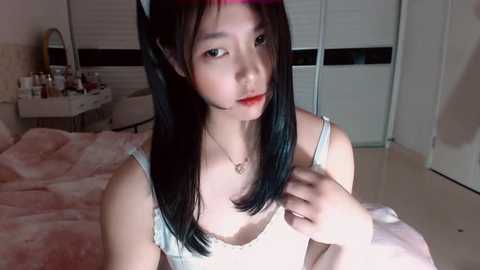 Video of a young Asian woman with long black hair, wearing a white lace bra, in a dimly lit bedroom with a pink bedspread and closed blinds.