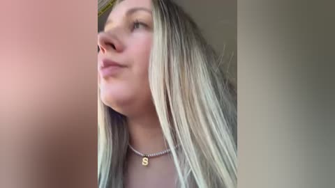 Video of a young Caucasian woman with long, straight blonde hair, fair skin, and a slender figure. She wears a simple gold necklace. The background is out of focus, blending from a warm pink to a soft beige.