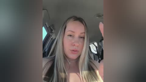 Video of a blonde woman with fair skin and medium build, smiling, sitting in a car. She has long, straight hair, wearing a choker necklace. The car's interior is visible, with a sunroof and dark seats.