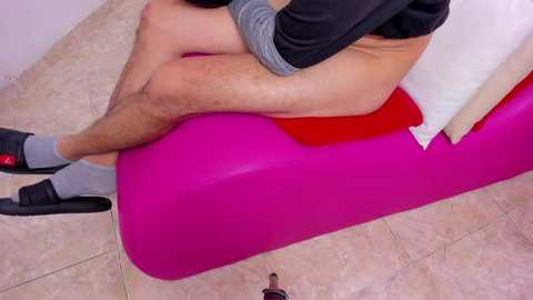Video of a person with fair skin, wearing grey socks and black shoes, sitting on a pink inflatable raft with white cushion. The background features a beige tiled floor.