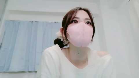 A video of an East Asian woman with a pale complexion and brown hair tied back, wearing a pink face mask and a white shirt. She is in a sterile, white-tiled bathroom with a light blue curtain in the background.