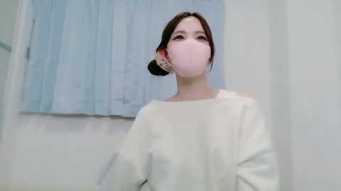 Video of a woman in a white mask and white shirt, standing in a minimalist room with light blue curtains.