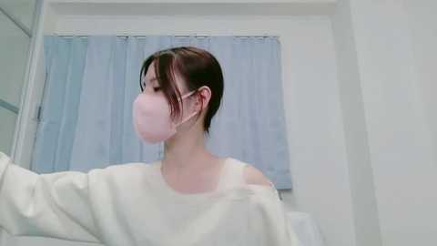 Video of a young East Asian woman with short dark hair, wearing a white off-shoulder sweater and a pink face mask, standing in a minimalist, brightly lit room with white walls and light blue curtains.