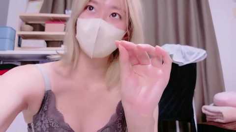 Video of a young Asian woman with fair skin, blonde hair, wearing a grey lace bra and a beige face mask, holding a white mask near her face, in a cluttered, possibly home office setting.