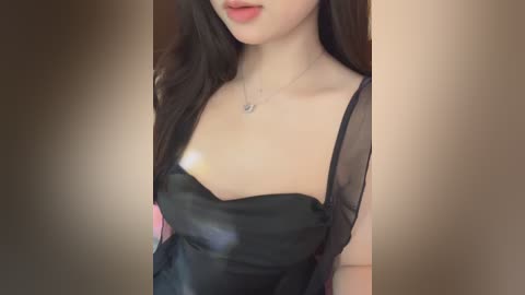 A video of a young Asian woman with long black hair, wearing a black, sheer dress, with a small heart pendant necklace, set against a blurred, beige background.
