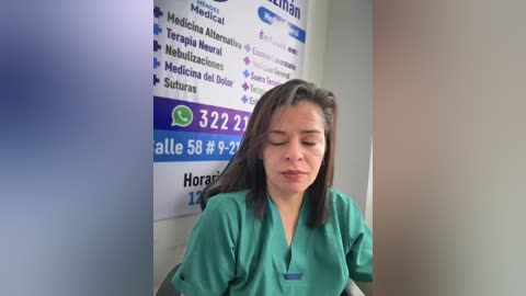 Video of a Latina woman with long dark hair in teal scrubs, sitting with eyes closed, in a medical office with white walls and blue informational posters.