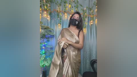 Video of a woman in a gold sari with a black mask, standing in a room with green curtains, fairy lights, and a potted plant.