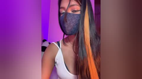 Video of a young woman with medium skin tone, long dark hair, wearing a black face mask, white tank top, and multicolored striped socks. Background is a purple-lit room.