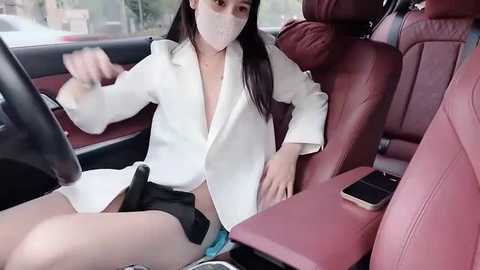 Video of an Asian woman with long black hair, wearing a white lab coat and mask, sitting in a car with red leather seats, driving with a black steering wheel.