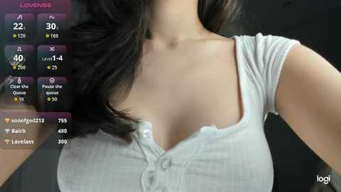 A video of a woman with fair skin and dark hair, wearing a tight, light gray, short-sleeved, button-down shirt. The background is dark and blurred. Text overlays show social media metrics.