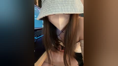 Video of a young woman with long brown hair, wearing a light gray bucket hat and a white spaghetti strap top, sitting at a computer desk.
